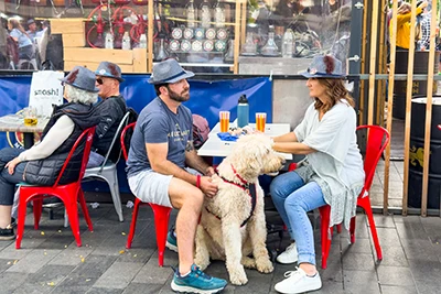 a young couple wearing the symbolic grey hat of the Oktoberfest sitting at an outdoor table with theri large,offwhite dog sitting between them