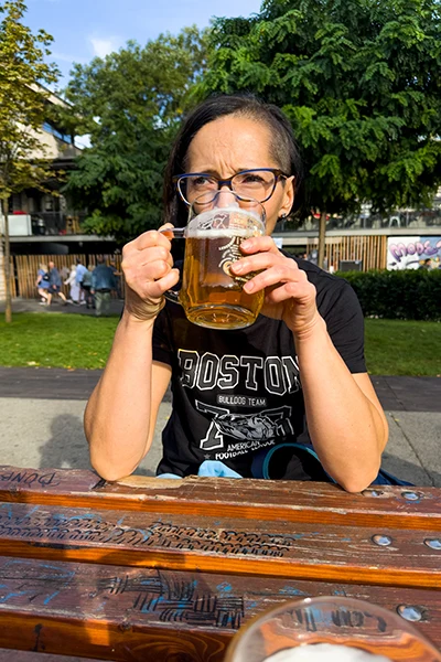 middle-aged woman with dark hair and wearing glasses drinking a mug of wheat beer sitting a wooden bench in apark