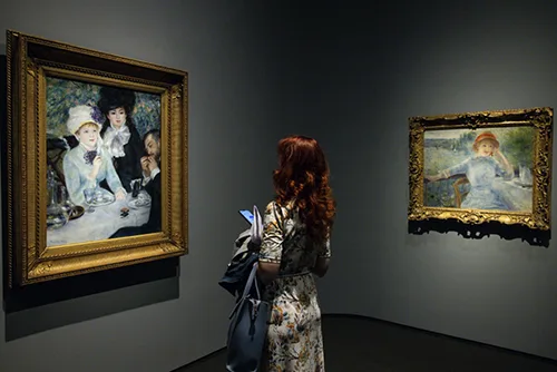 a woman with long auburn hair looking at two of the exhibited paintings of the Renoir Exhibition in the Hungarian Museum of Fine Arts,