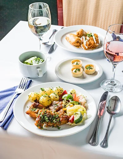 Roast chicken in gravy with boiled potatoes and steamed veggies, stuffed cabbage - both dishes served on round white plates, a glass of white and rose wine on the white clothed table