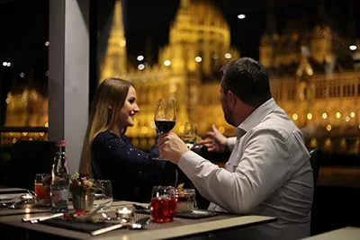 a couple drinking red wine possibly on a Danube cruise, the lit parliament can be seen through the window in the background