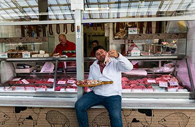 a butcher sitting on the sounter of his shop and holding a wooden board packed with cold cuts and meats