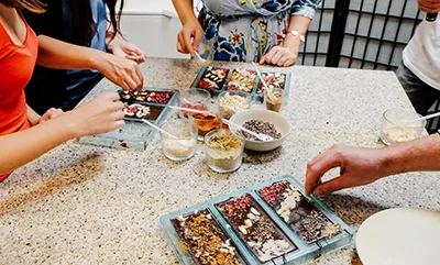 cc.five people around a marble top creating their own chocolate bars, adding nuts and other ingredients
