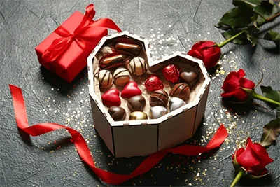 bonbons in a heart-spabed box with three red roses on its left and a red ribbon