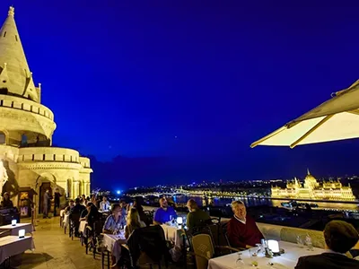 Diners sitting at tables on the panorama terrace of the Halaszbastya Restaurant at the blue hour. View of the lit up Parliament in Pest si visible
