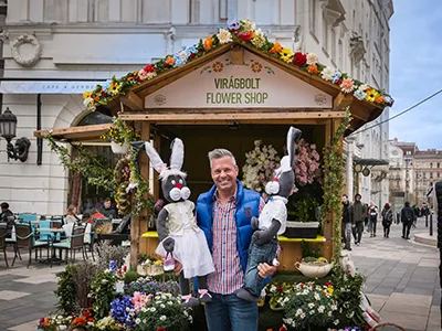 a man holding two stuffed Easter bunnies in his hands, he stands in front of a lovely flower shop booth - Vorosmarty Square Easter market