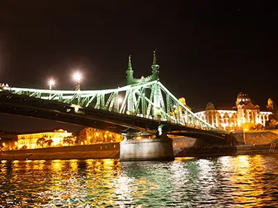 the green Liberty Bridge illuminated at night as seen from aboat on the Danube, the Gellert Bath is visible in the background