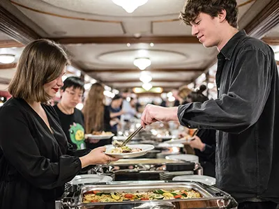 a young man and woman (both dressed in black) selecting food from the buffet dinner table on a ship