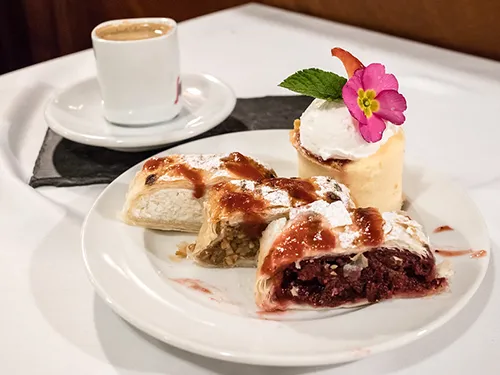 3 pieces of strudel (cottage cheese, apple, sour cherry filling) on a round white dessert plate, a cup of coffee next to it