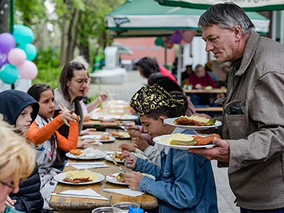 an older man holding two plates of food in his hands, probbaly about to sit down to eat , a handful of other people already sitting and eating at a wooden table