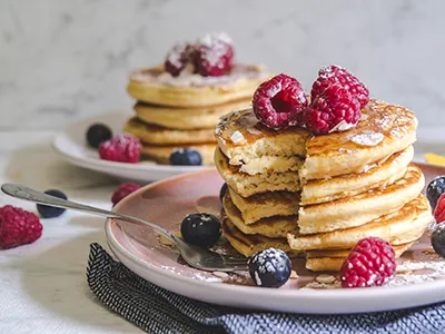 a stack of American pancakes with a a couple of raspberries and blueberries on a round pale pink plate