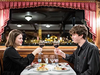 a young woman and man raising their glaases of red wine to toast, they're having dinner on board the Szechenyi cruise boat